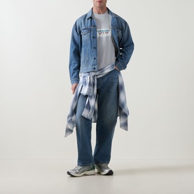 Model wearing Carhartt Bumper Stickers Logo T-Shirt in Pontiki grey, styled with denim jacket, wide-leg jeans and a tied checked shirt at waist.