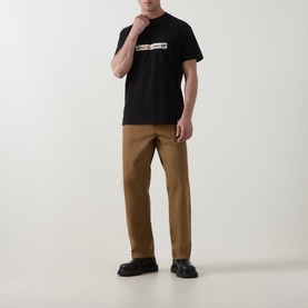Model wearing Carhartt Bumper Stickers Logo T-Shirt in black with graphic chest print, styled with tan trousers and black chunky shoes.