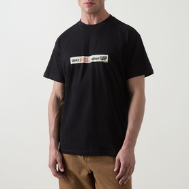 Model wearing Carhartt Bumper Stickers Logo T-Shirt in black, crew neck with graphic print, paired with tan trousers
