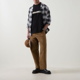 Model wearing Carhartt Bumper Stickers Logo T-Shirt in Black, styled with plaid overshirt, tan trousers and black shoes.