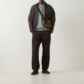 Model wearing Carhartt Caspian Sweatshirt in Shady Green Heather with dark trousers and jacket, styled with a tan crossbody bag.