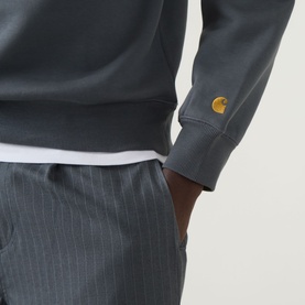 Model wearing Carhartt Chase Logo Sweatshirt in Styx/Gold, showing gold embroidered logo above cuff, styled with grey pinstripe trousers.