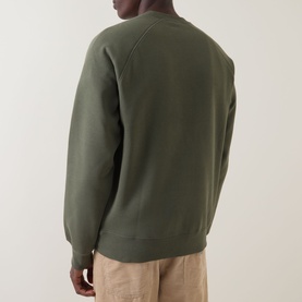 Model wearing Carhartt Chase Logo Sweatshirt in Leaf/Gold. Rear view showing crew neck, raglan sleeves, and ribbed hem. Styled with beige trousers.