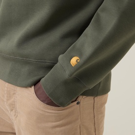 Model wearing Carhartt Chase Logo Sweatshirt in Leaf/Gold, close-up of embroidered gold logo above ribbed cuff, styled with beige trousers.