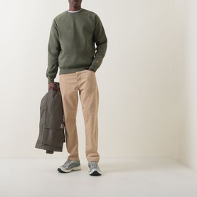 Model wearing Carhartt Chase Logo Sweatshirt in Leaf/Gold with beige trousers, carrying a brown jacket, styled with grey trainers.