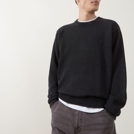 Model wearing Carhartt Kori Logo Knit Sweater in black, crew neck with ribbed trims and tonal chest logo, layered over white tee with grey trousers.