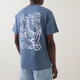 Model wearing Carhartt Formidable Logo T-Shirt in Shady Blue, showing back with enlarged white tiger graphic print, styled with light-wash jeans.