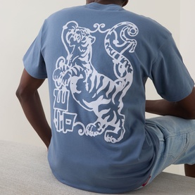 Model wearing Carhartt Formidable Logo T-Shirt in Shady Blue, showing enlarged white tiger graphic print on back, styled with light blue jeans.