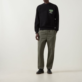 Model wearing Carhartt Pinball Logo Sweatshirt in black with chest graphic, styled with khaki trousers and black loafers.