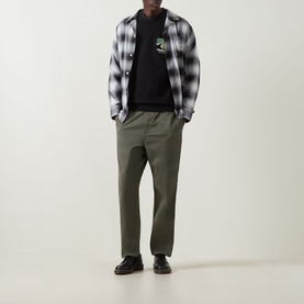 Model wearing Carhartt Pinball Logo Sweatshirt in Black under grey plaid overshirt, styled with olive trousers and black leather shoes.