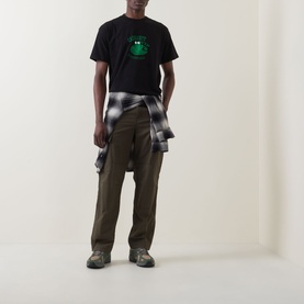 Model wearing Carhartt Please Don't Snitch Logo T-Shirt in black with green frog graphic, styled with olive trousers and plaid shirt tied at waist.