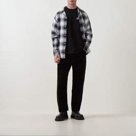 Model wearing Carhartt Pup Logo T-Shirt in Black under grey plaid overshirt, styled with black trousers and chunky black loafers.