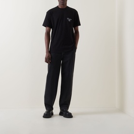 Model wearing Carhartt Gracious Logo T-Shirt in black with chest embroidery, styled with black wide-leg trousers and chunky loafers.