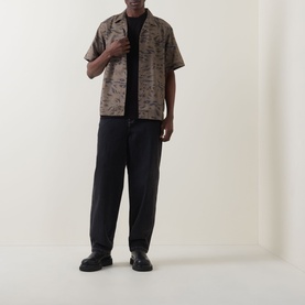 Model wearing Carhartt Gracious Logo T-Shirt in Black, styled with a brown printed short-sleeve shirt, black trousers and chunky loafers.