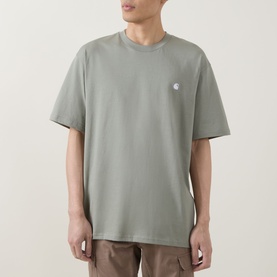 Model wearing Carhartt Madison Logo T-Shirt in Shady Green with white embroidered chest logo, crew neck, short sleeves, paired with brown trousers.