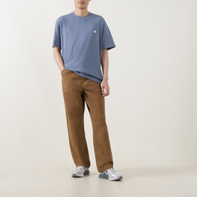Model wearing Carhartt Crew Neck T in Shady Blue with small chest logo, styled with tan trousers and grey trainers.
