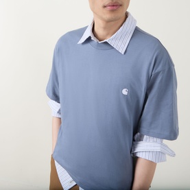 Model wearing Carhartt Crew Neck T in Shady Blue with small chest logo, styled over a blue striped shirt with spread collar.