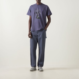 Model wearing Carhartt Fan Graphic Logo T-Shirt in Shady Purple with straight hem, styled with blue trousers and grey trainers.