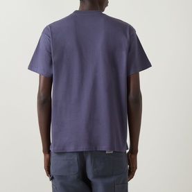 Model wearing Carhartt Fan Graphic Logo T-Shirt in Shady Purple, rear view, crew neck, short sleeves, straight hem, styled with grey trousers.