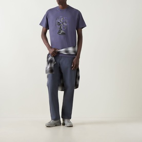 Model wearing Carhartt Fan Graphic Logo T-Shirt in Shady Purple with grey trousers and plaid shirt tied at waist.