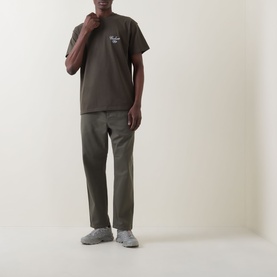 Model wearing Carhartt Gracious Logo T-Shirt in Cypress brown with chest embroidery, styled with khaki trousers and grey trainers.