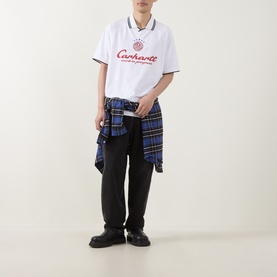 Model wearing Carhartt Heart Train/White polo-style T-shirt with red logo, styled with black trousers and blue tartan shirt tied at waist.