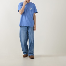 Model wearing Carhartt Waves Logo T-Shirt in Curacao blue with chest graphic, styled with light wash wide-leg jeans and brown suede shoes.