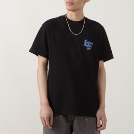 Model wearing Carhartt Waves Logo T-Shirt in black with blue chest graphic, styled with grey trousers and silver chain necklace.