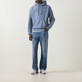 Model wearing Carhartt American Script Logo Hoodie in Shady Blue with blue mid-wash jeans and grey trainers.