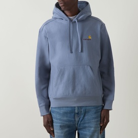 Model wearing Carhartt American Script Logo Hoodie in Shady Blue with kangaroo pocket and gold embroidered chest logo, styled with blue jeans