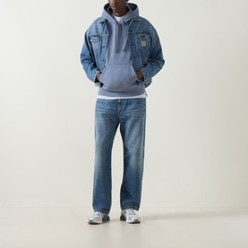 Model wearing Carhartt American Script Logo Hoodie in Shady Blue, styled with a denim jacket, light blue jeans, and grey trainers.