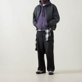 Model wearing Carhartt American Script Logo Hoodie in Shady Purple, styled with dark bomber jacket, check shirt, and black wide-leg trousers.