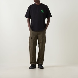 Model wearing Carhartt Built Different Logo T-Shirt in black with green chest graphic, styled with olive cargo trousers and black shoes.