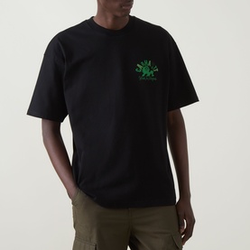 Model wearing Carhartt Built Different Logo T-Shirt in black with green chest graphic, crew neck, dropped shoulders, short sleeves, styled with khaki