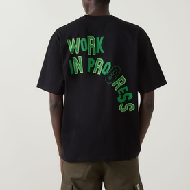 Model wearing Carhartt Built Different Logo T-Shirt in Black, showing rear with green Work In Progress graphic print, styled with khaki trousers.