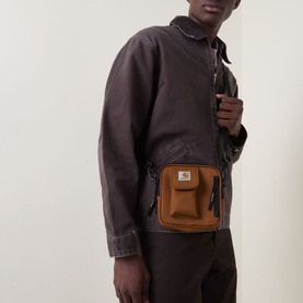 Model carrying Carhartt Essentials Logo Crossbody Bag in Hamilton Brown, worn across chest with adjustable strap, styled with dark work jacket