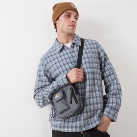 Model carrying Carhartt Essentials Logo Crossbody Bag in Styx grey, worn crossbody with black strap, styled with plaid shirt and tan beanie.