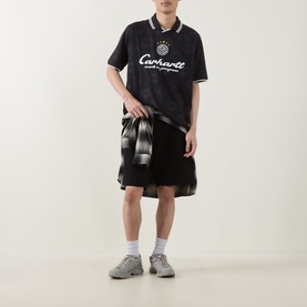Model wearing Carhartt Heart Train polo shirt in black with white logo print, styled with black check shirt tied at waist and black shorts.