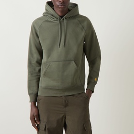 Model wearing Carhartt Chase Logo Hoodie in Leaf/Gold, styled with khaki cargo shorts. Kangaroo pocket and gold embroidered cuff logo visible.