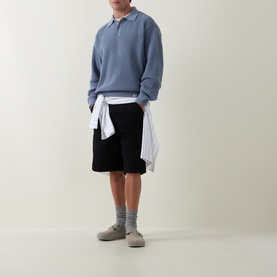 Model wearing Carhartt Shady Blue cotton polo knit, styled with white shirt tied at waist, black shorts and grey shoes