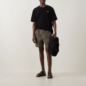 Model wearing Carhartt shorts in Duck Feather camo print, styled with black T-shirt and sandals, carrying a black bag.