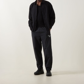Model wearing Carhartt Travon Script Logo Track Pants in Black, styled with a black bomber jacket and black trainers.