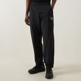 Model wearing Carhartt Travon Script Logo Track Pants in Black, straight-leg fit with embroidered logo, styled with black chunky trainers.
