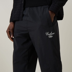 Model wearing Carhartt Travon Script Logo Track Pants in black, showing embroidered script logo detail and side zip pocket on straight-leg fit.