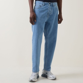 Model wearing Carhartt Newel Tapered Leg Jeans in blue, relaxed tapered fit with five-pocket styling, paired with grey trainers.