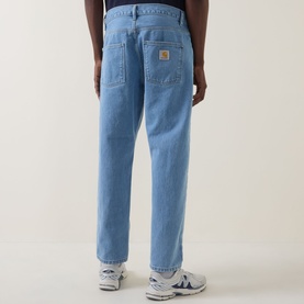 Model wearing Carhartt Newel Tapered Leg Jeans in blue, rear view showing woven logo badge and relaxed tapered fit, styled with white trainers.