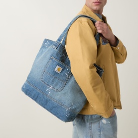 Model carrying Carhartt Logo Denim Tote Bag in mid-wash blue denim with front patch pocket, logo badge, and tobacco stitching, over shoulder.