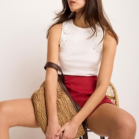 Model wearing Mint Velvet white sleeveless top with floral detail, styled with red mini skirt; model seated holding woven straw clutch bag.