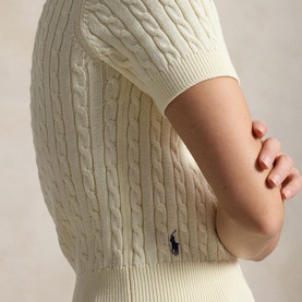 Model wearing Polo Ralph Lauren cable-knit jumper in Clubhouse Cream, with navy embroidered pony logo detail, arms crossed.