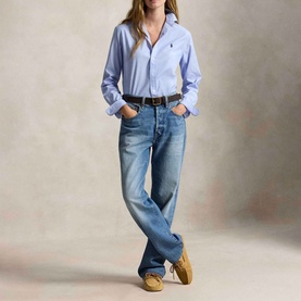 Model wearing a light blue long-sleeve shirt with navy logo, styled with wide-leg jeans, brown belt, and tan suede shoes.